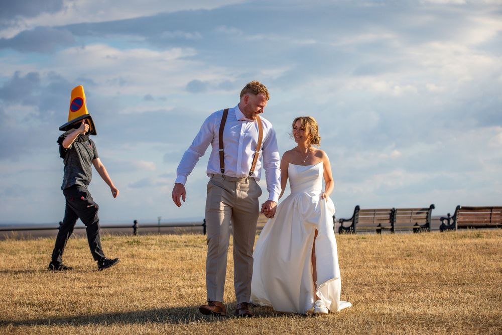 Bride and groom hold hands in a sunny field, with a person in a cone hat walking behind them.