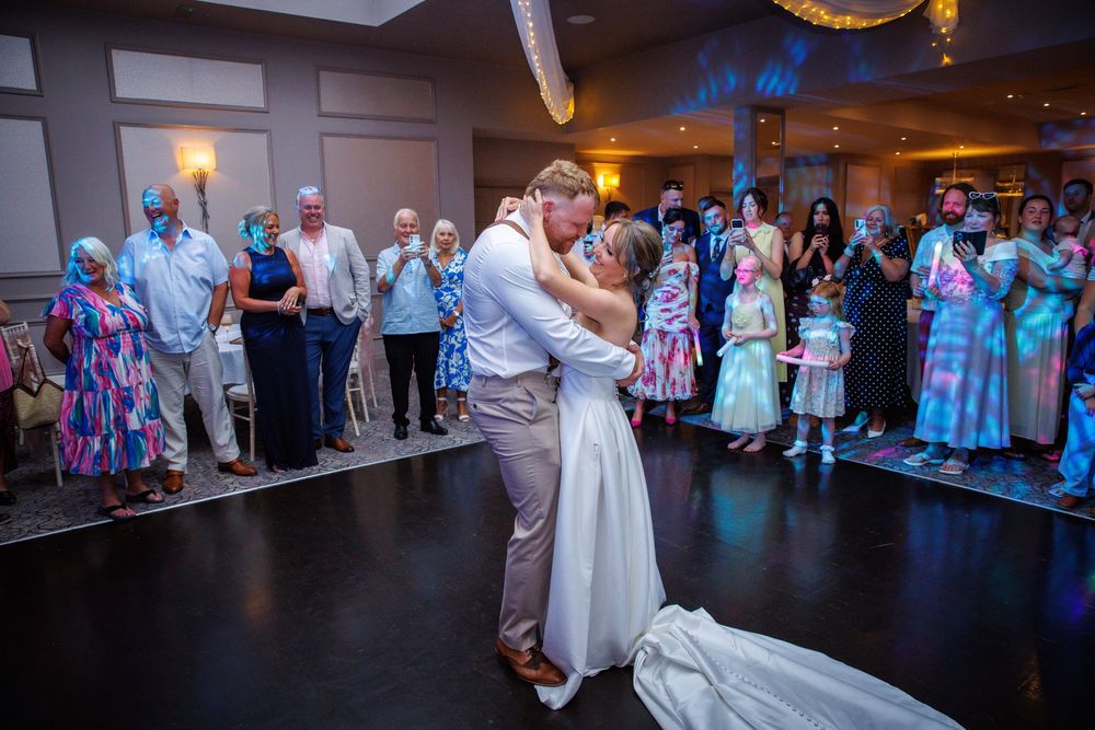 Bride and groom share first dance kiss at wedding reception while guests watch and celebrate around the dance floor.
