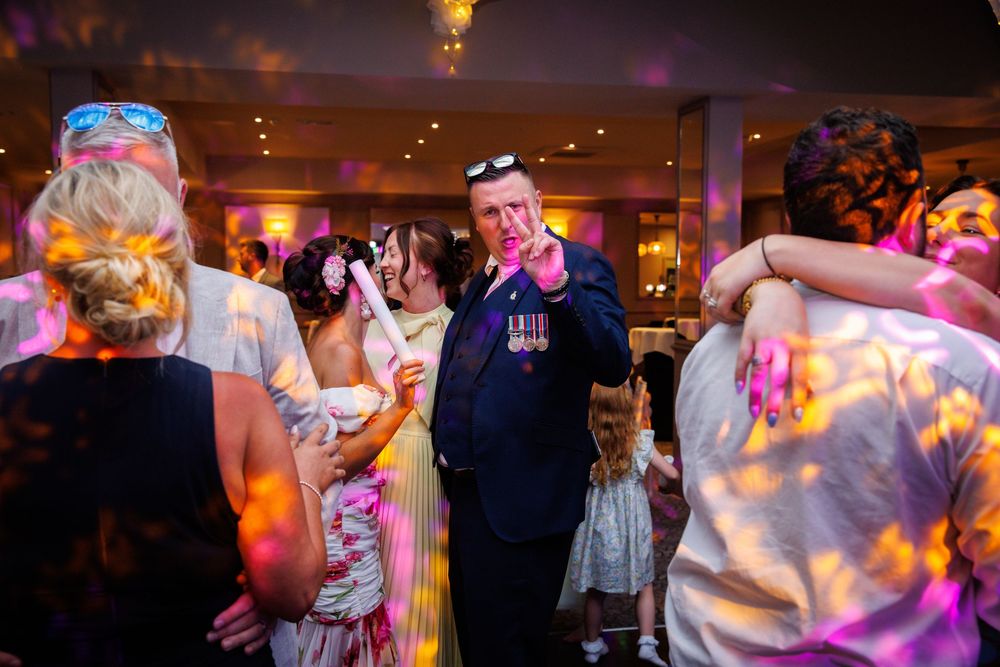 Guests dancing at a wedding reception with colorful disco lights, featuring a man in military uniform smiling.