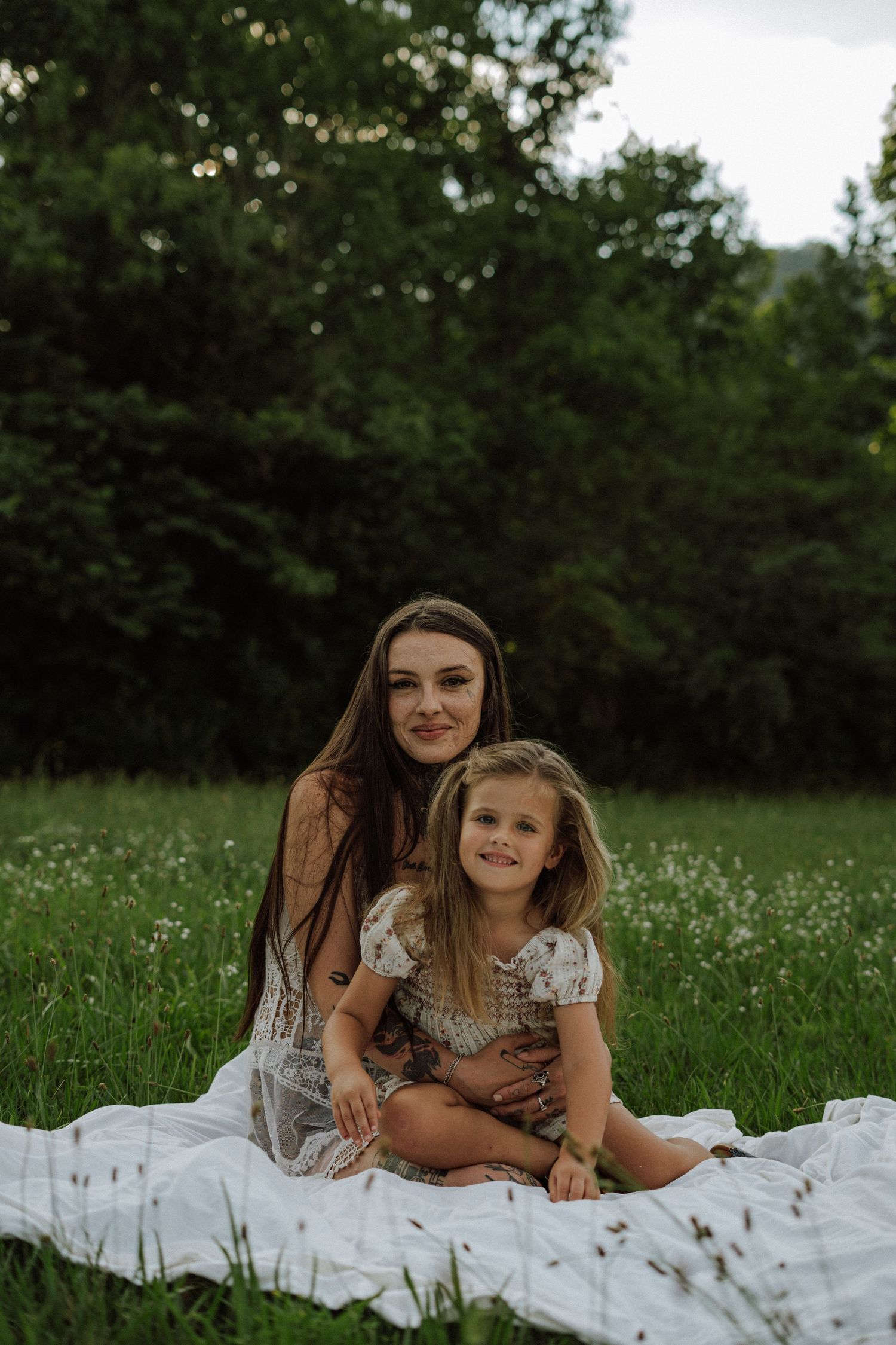 A mother and young daughter sitting together on a white blanket in a lush green meadow surrounded by trees.