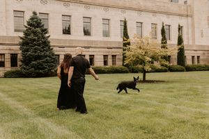 Couple walking with black dog on green lawn outside large stone building.