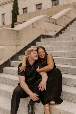 A happy couple in black outfits sitting together on stone steps, smiling and embracing outdoors.