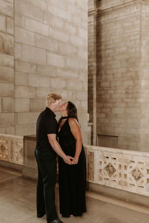 Couple in matching black outfits sharing a romantic kiss in front of elegant stone architecture columns.
