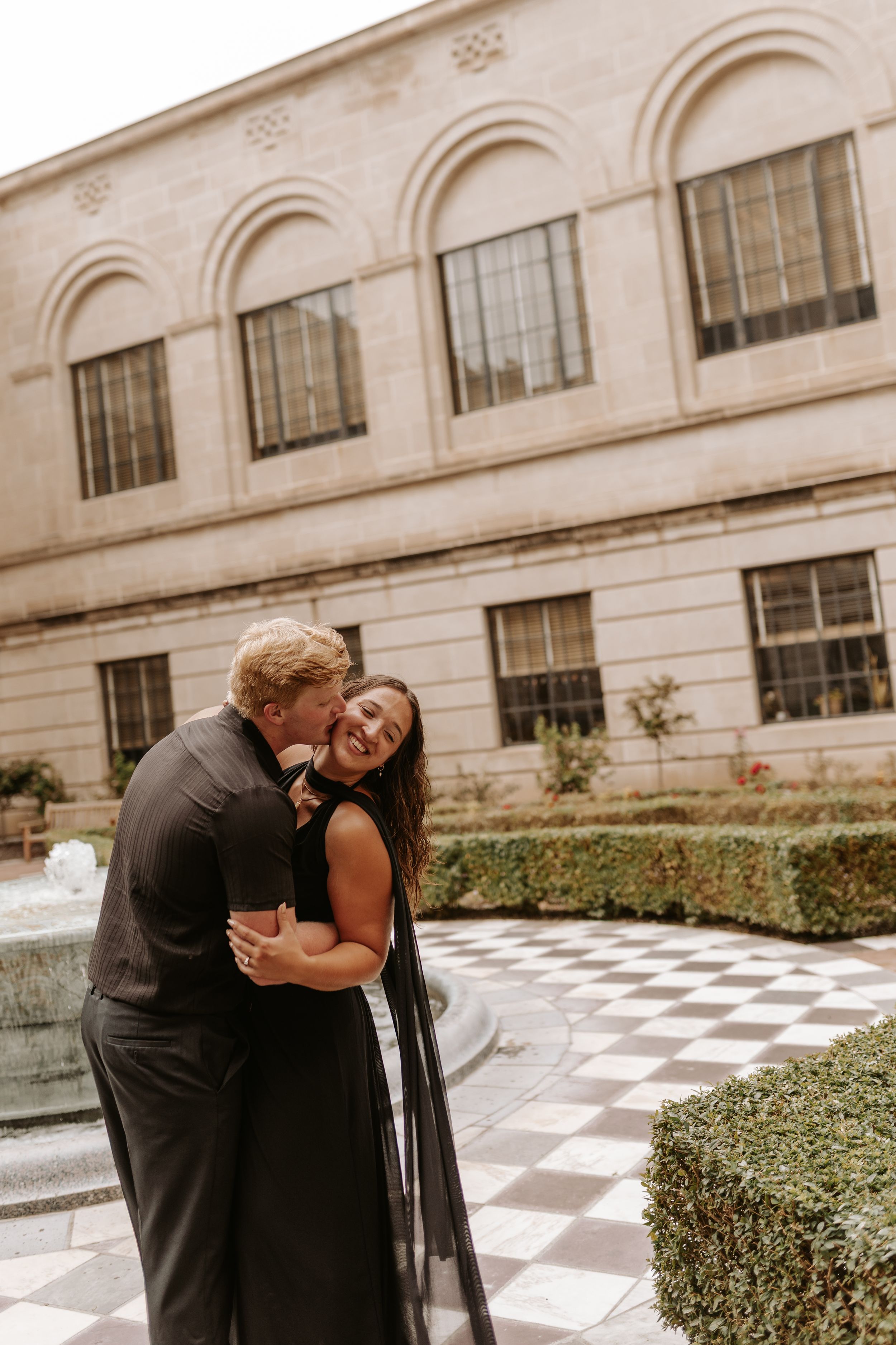 Couple embracing and laughing in elegant black attire outside a classic stone building with arched windows.