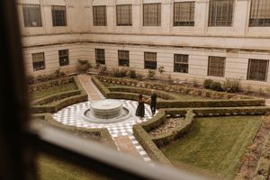 Couple standing in a formal garden courtyard with geometric hedges and a fountain, viewed from above.