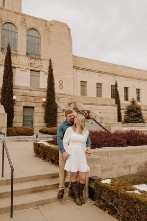 Couple holding hands and smiling on stone steps outside a classic limestone building with cypress trees.