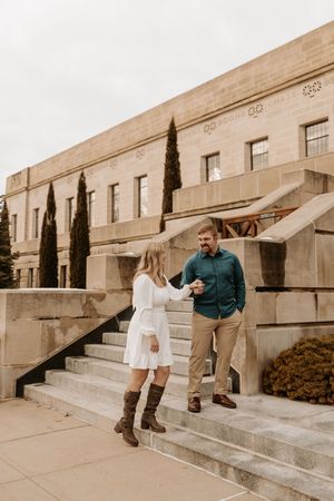 Couple holding hands on stone steps outside a classic building, woman in white dress and boots, man in teal shirt.