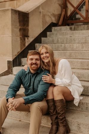 Smiling couple sitting on stone steps, woman in white dress and brown boots embracing red-haired man.