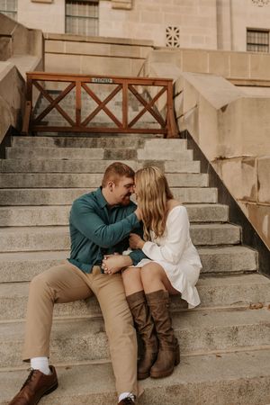 Couple sitting close on stone steps, sharing a romantic moment, man in teal shirt and woman in white dress with brown boots.