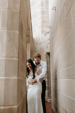 A laughing bride and groom embrace in a narrow stone alleyway, sharing a joyful moment on their wedding day.