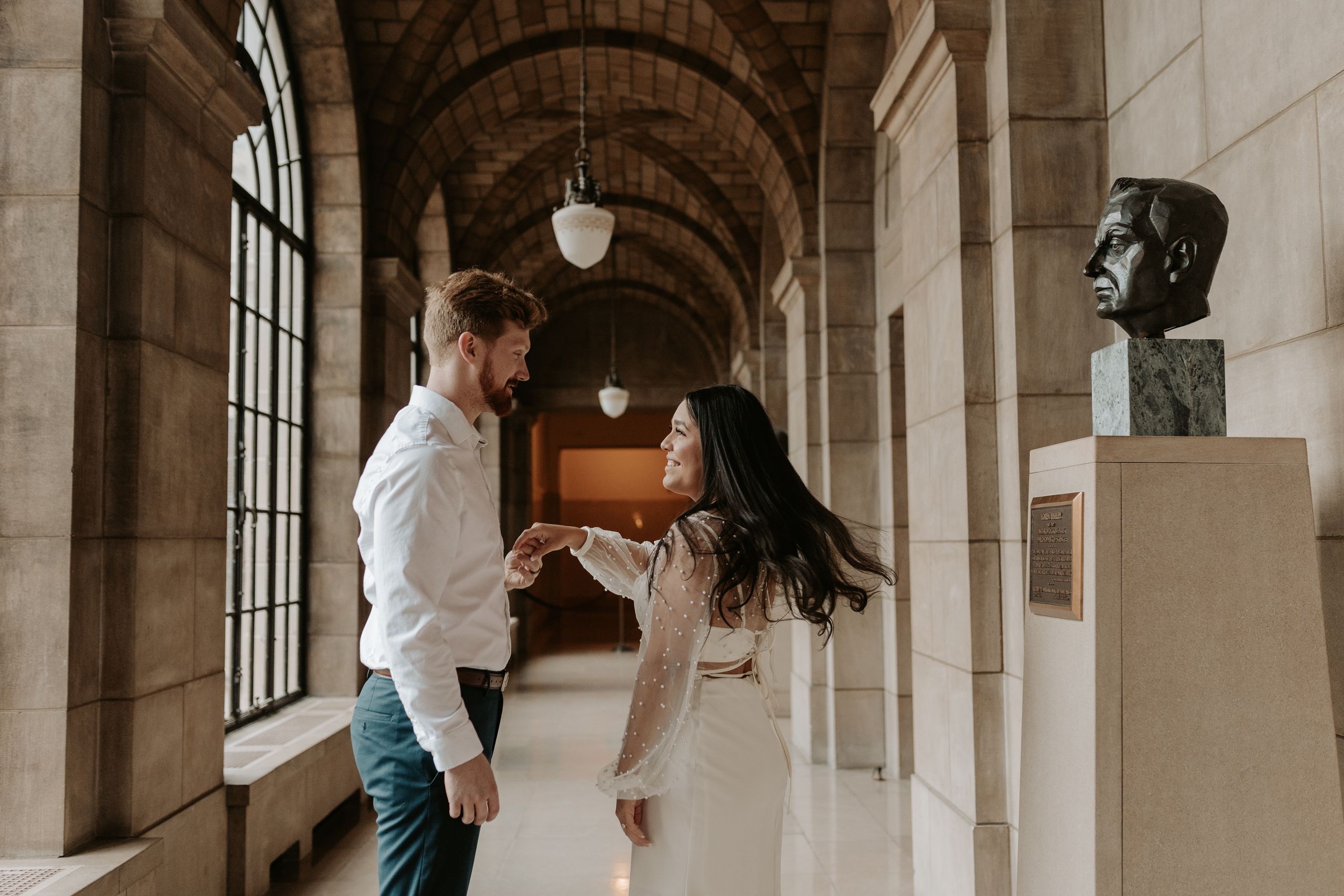 Couple dancing and holding hands in an elegant arched hallway with stone columns and a bronze bust sculpture.