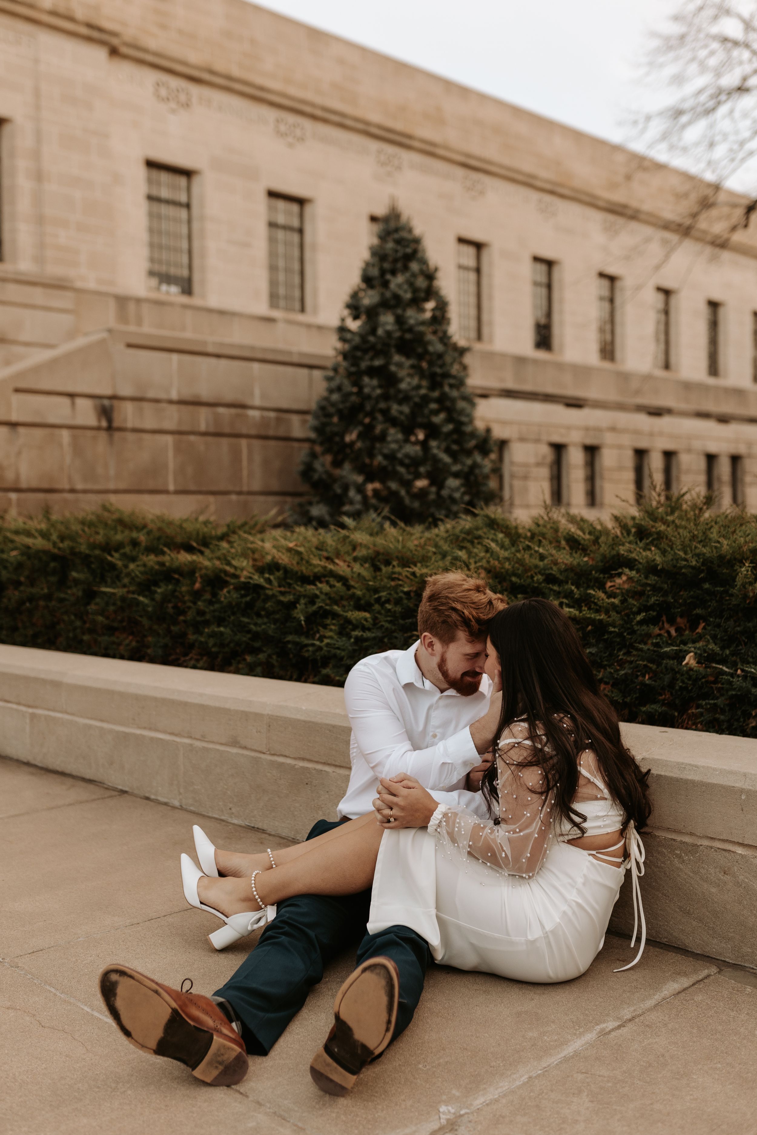 Couple sitting on stone steps in white outfits, sharing a romantic moment near a Christmas tree outside a classic building.