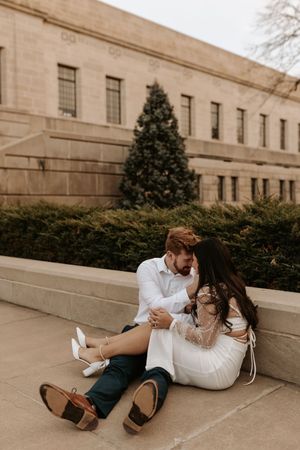Couple sitting on stone steps in white outfits, sharing a romantic moment near a Christmas tree outside a classic building.