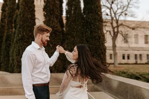 Couple dancing outdoors near cypress trees and historic building, woman in lace dress, man in white shirt.