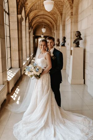 Bride and groom posing in elegant arched hallway, bride holding blue and white bouquet in lace gown.