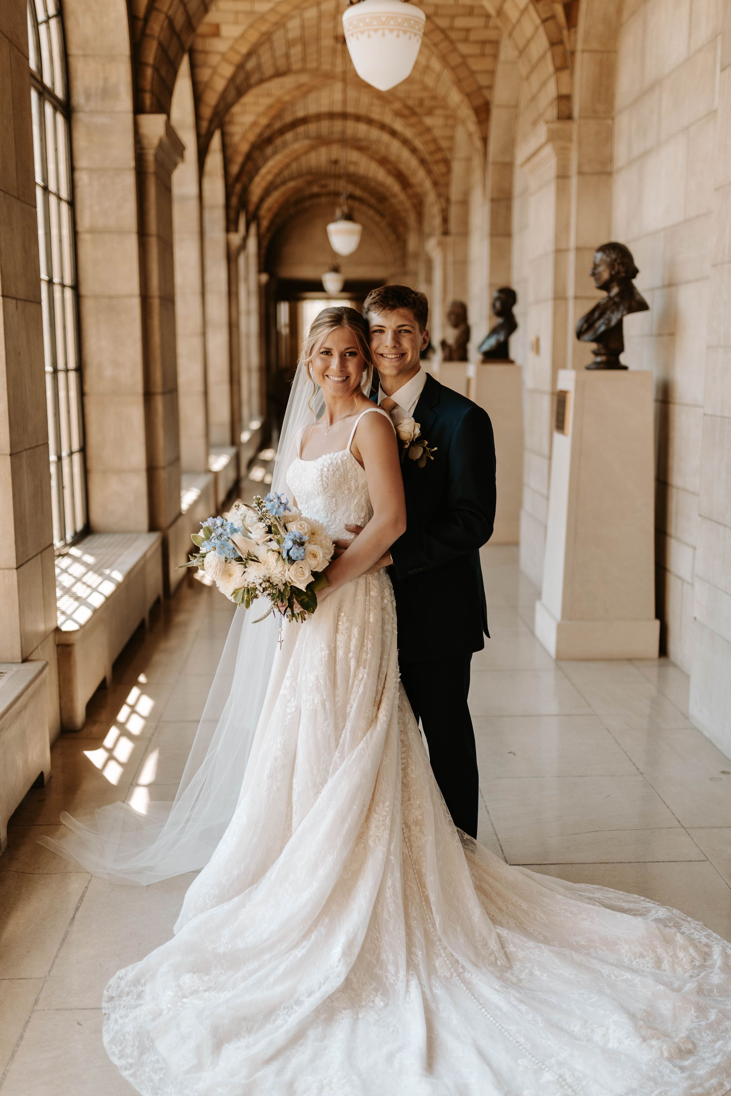 Bride and groom posing in elegant arched hallway, bride holding blue and white bouquet in lace gown.