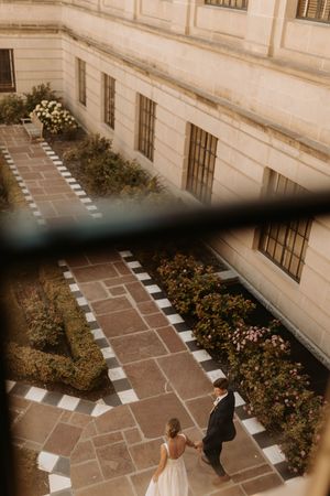 Aerial view of a wedding couple holding hands in an elegant courtyard garden with stone pathways and lush greenery.