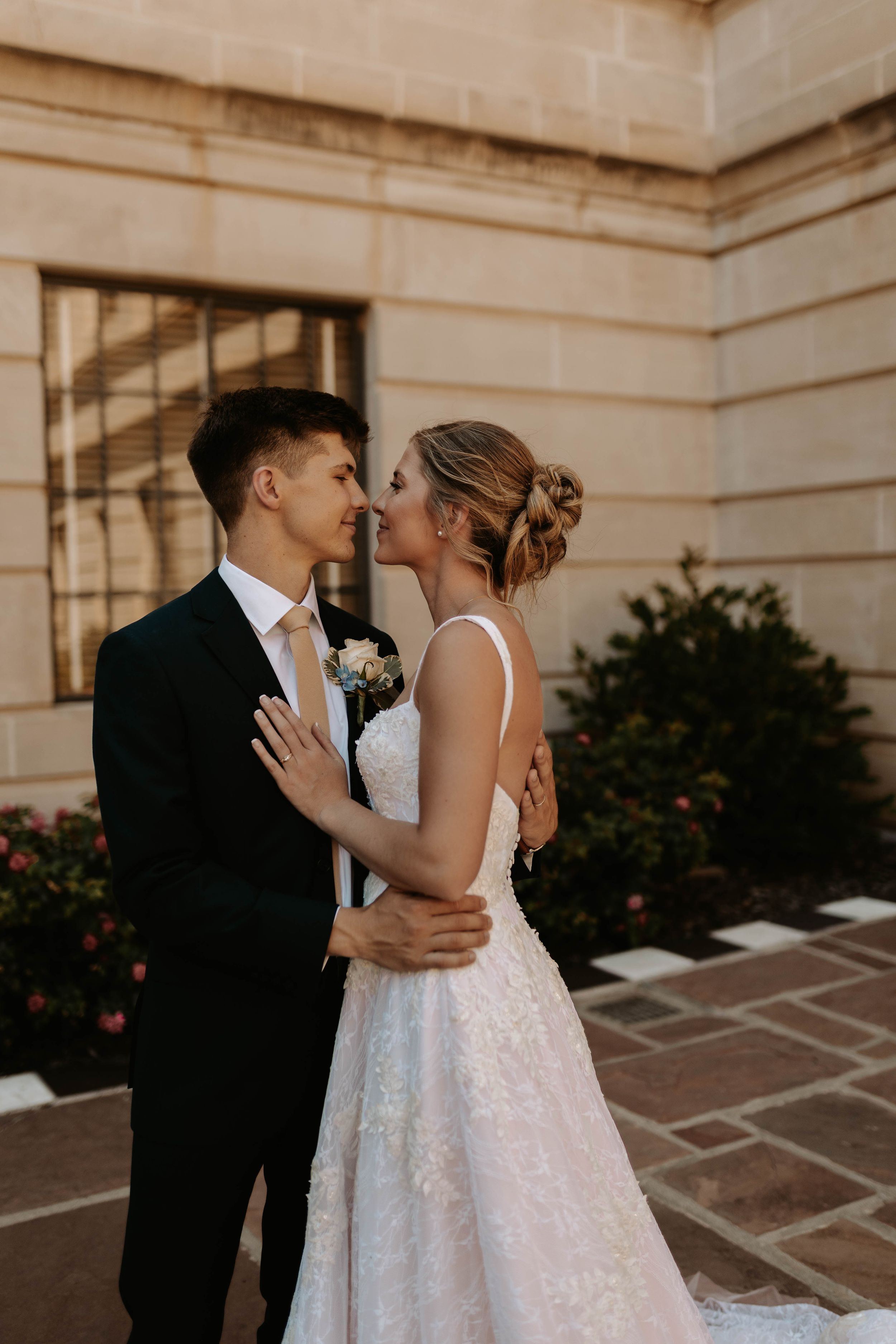 A bride and groom share a romantic embrace outside a stone building, with the bride in a lace gown and groom in a black suit.