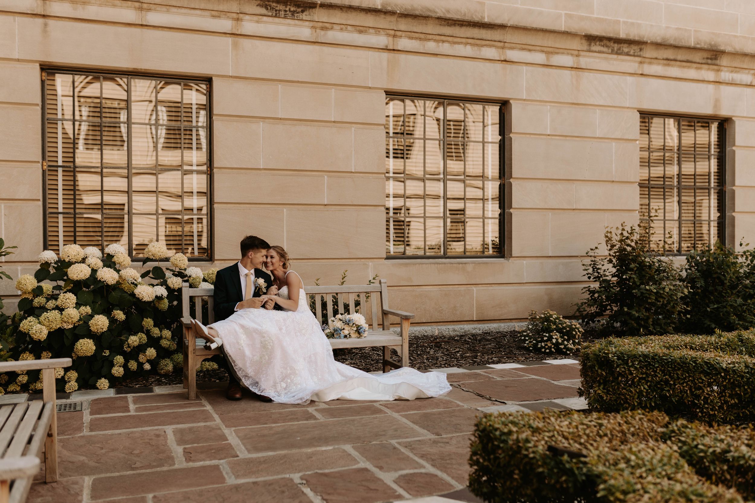 Bride and groom sitting together on a wooden bench in an elegant outdoor courtyard with white hydrangeas.
