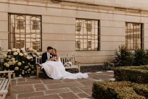 Bride and groom sitting together on a wooden bench in an elegant outdoor courtyard with white hydrangeas.