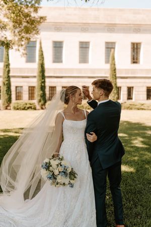 Bride and groom share a romantic moment outdoors, with veil flowing, bouquet of white and blue flowers, near elegant building.