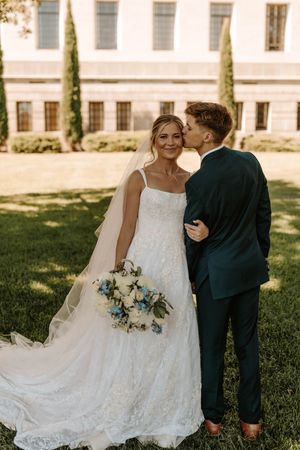 Bride in lace gown holding blue bouquet receives kiss from groom in navy suit outdoors near elegant building.