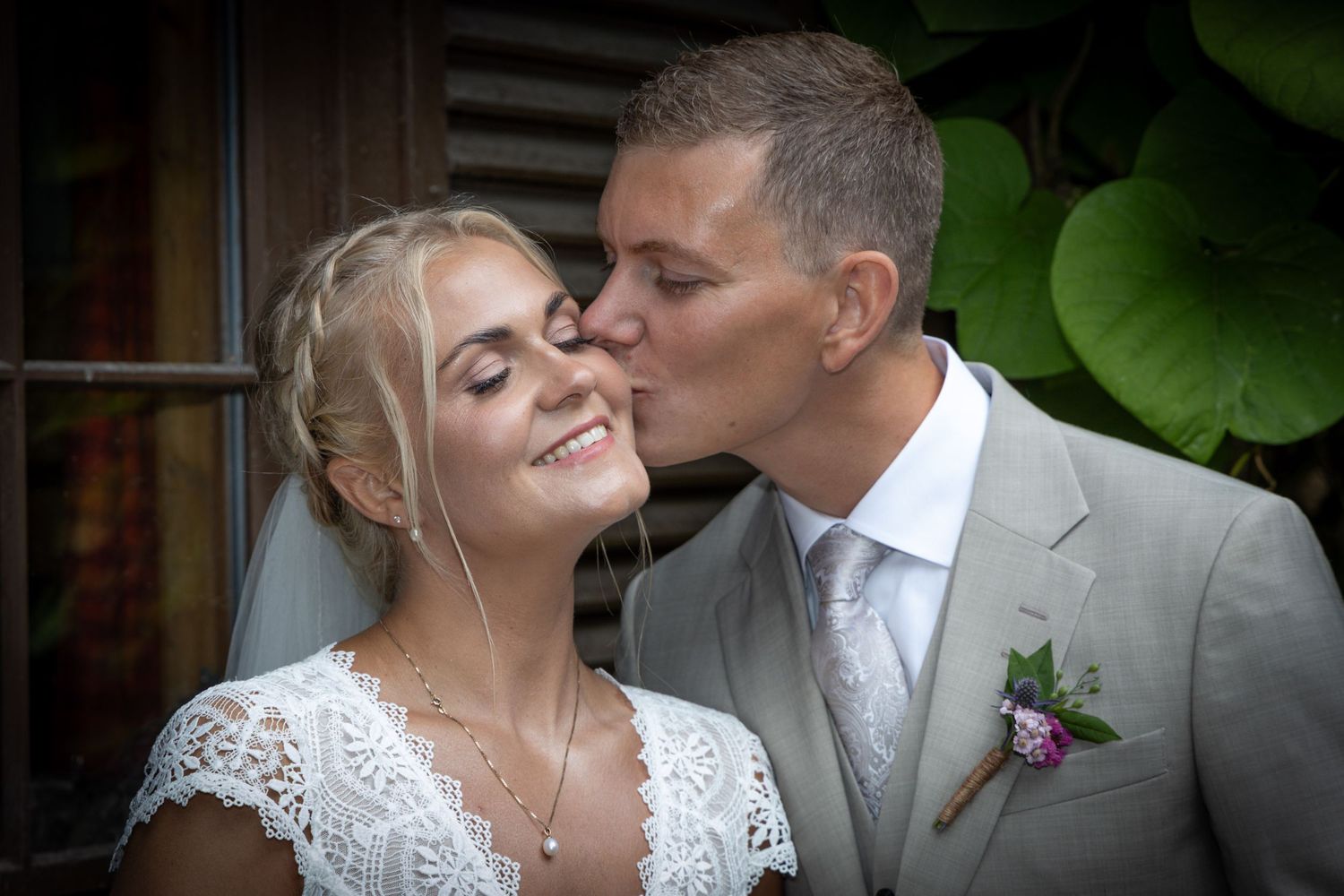 Groom in gray suit kisses bride in white lace dress on cheek as she smiles on their wedding day.