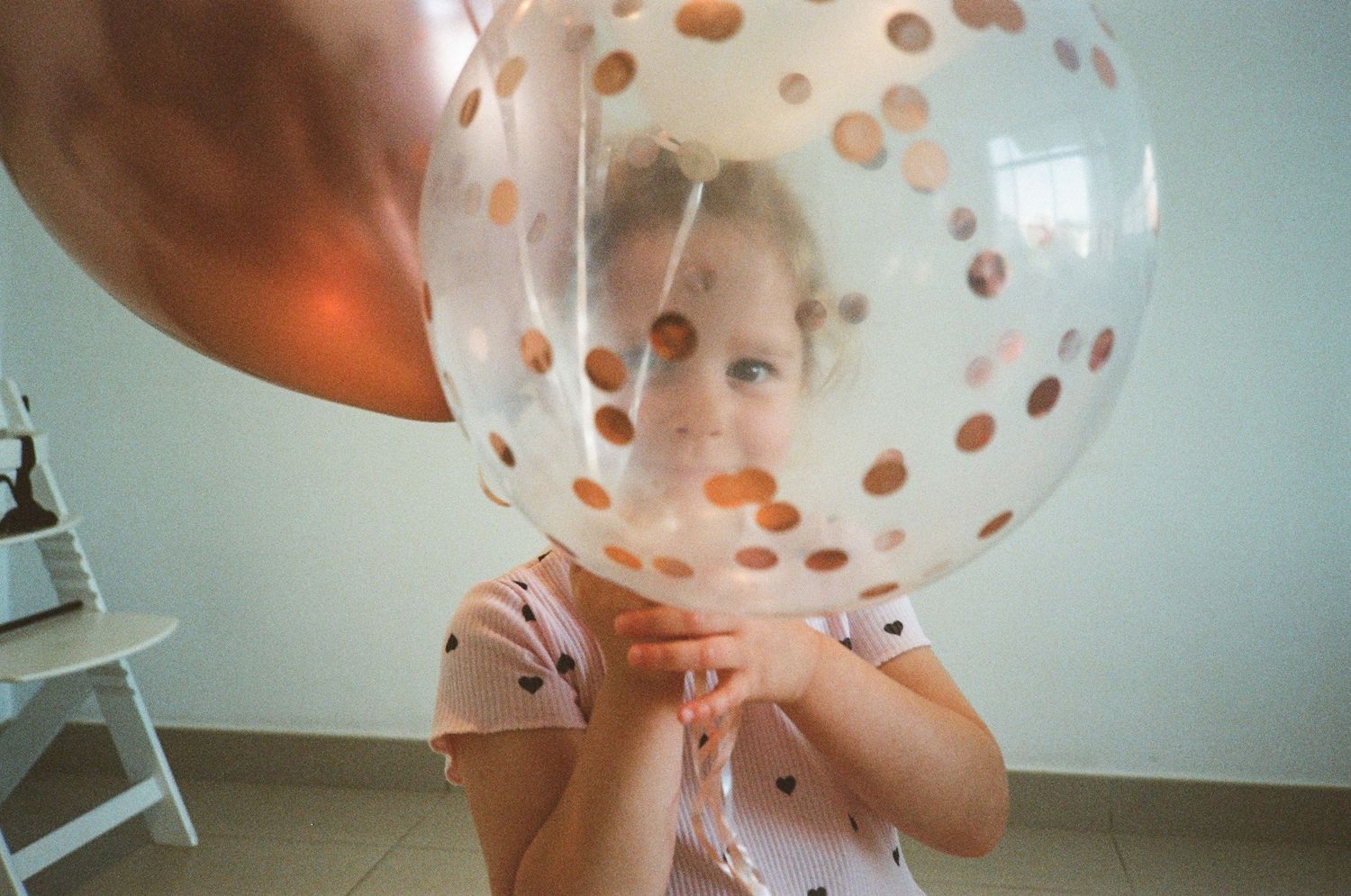 Young girl peering through a large clear confetti balloon at a birthday party celebration.