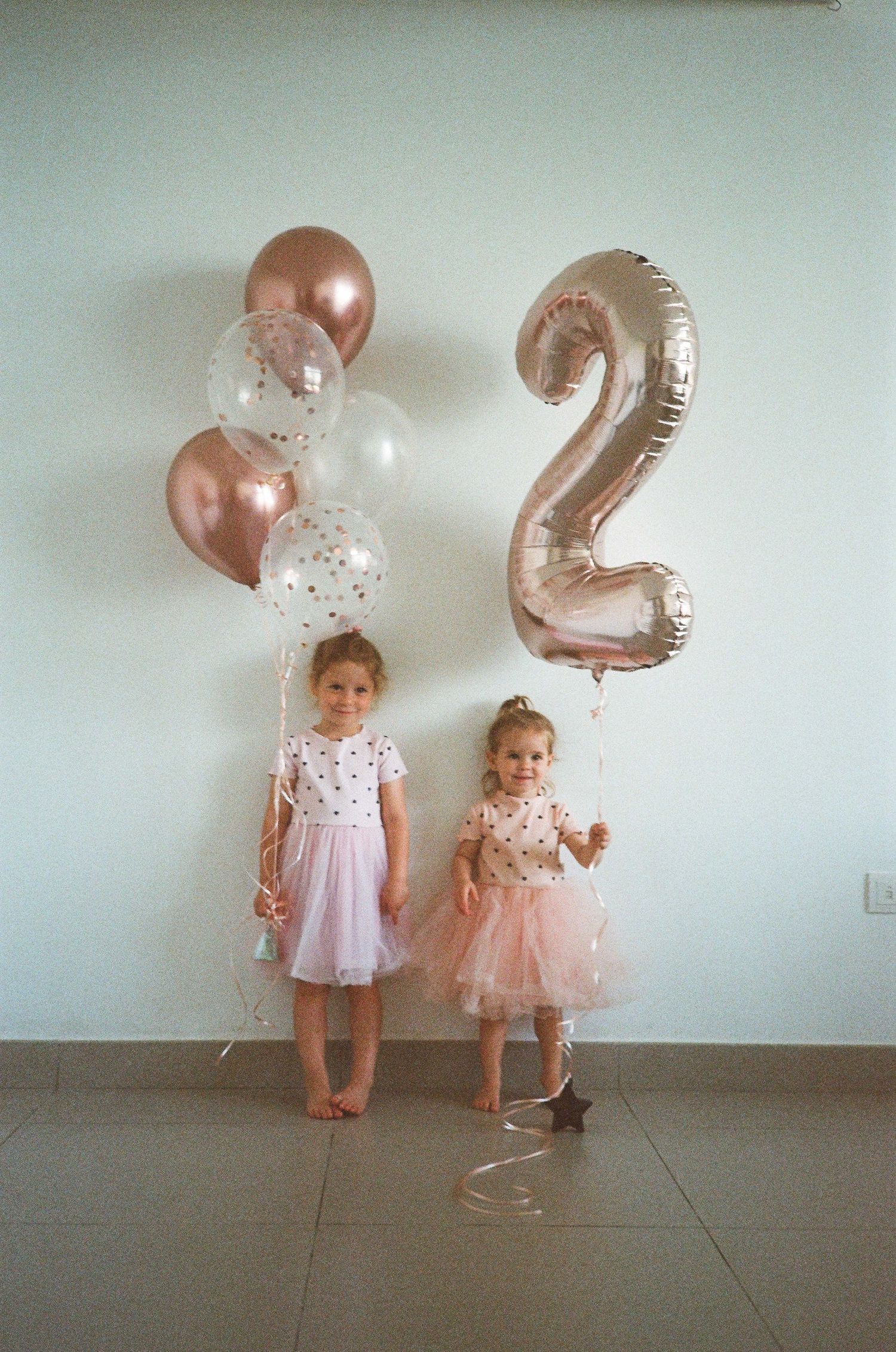 Two little girls in pink tutu dresses pose with rose gold balloons and a number 2 balloon at a birthday party.