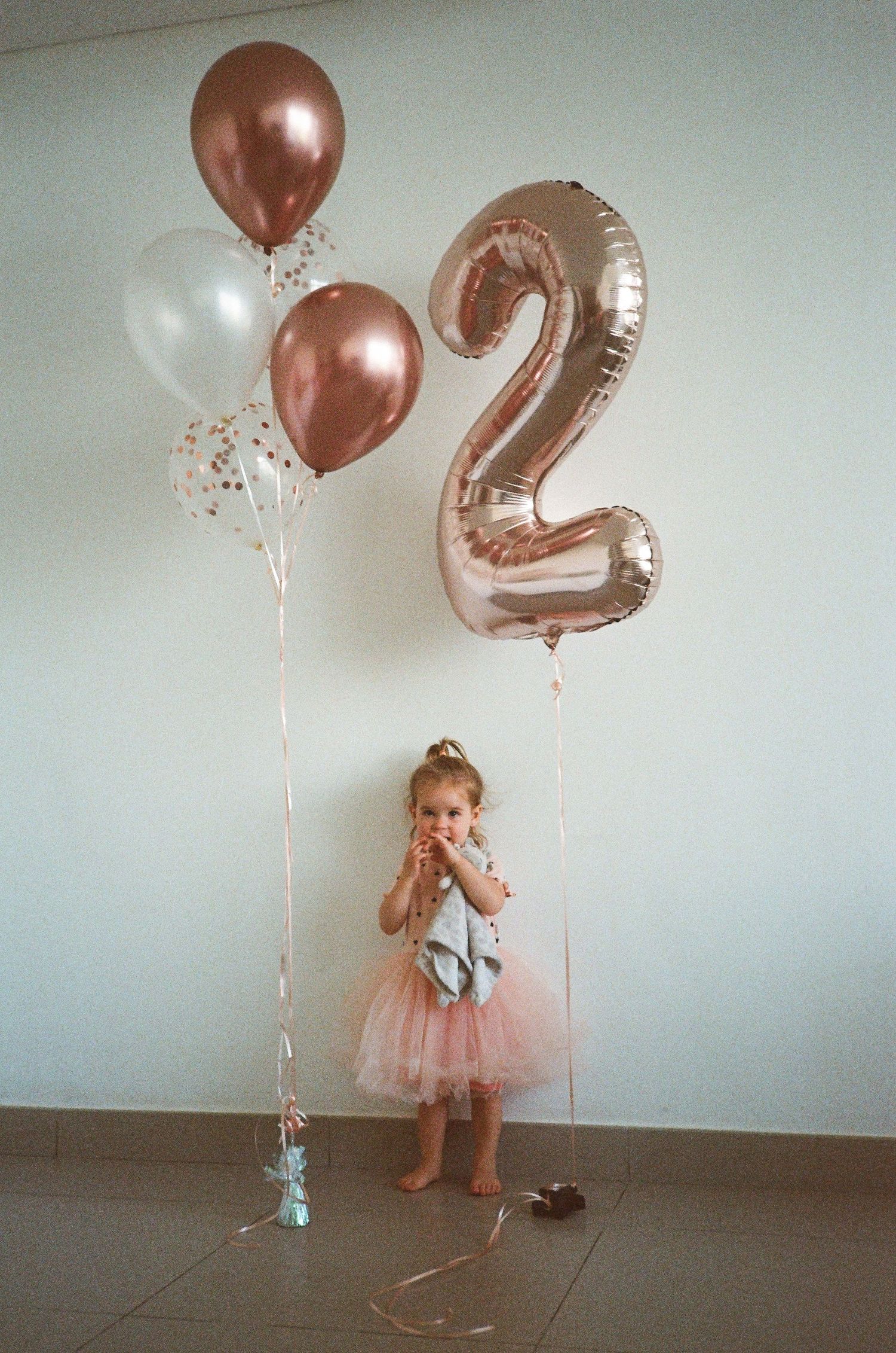 Toddler girl in pink tutu celebrates 2nd birthday with rose gold number 2 balloon and metallic balloons indoors.