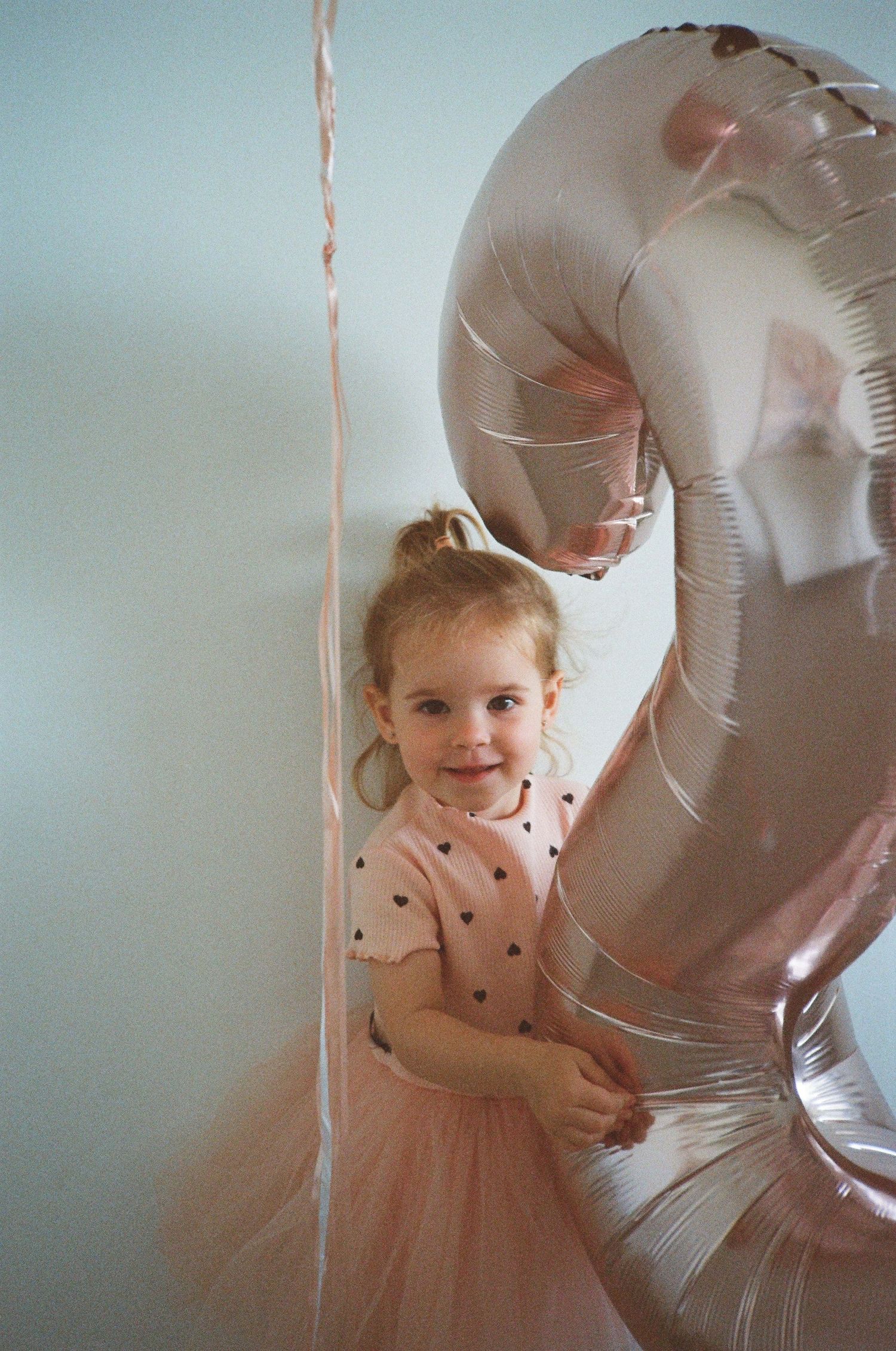 A smiling toddler girl in a pink tutu dress stands next to a large number 2 balloon at her birthday party.