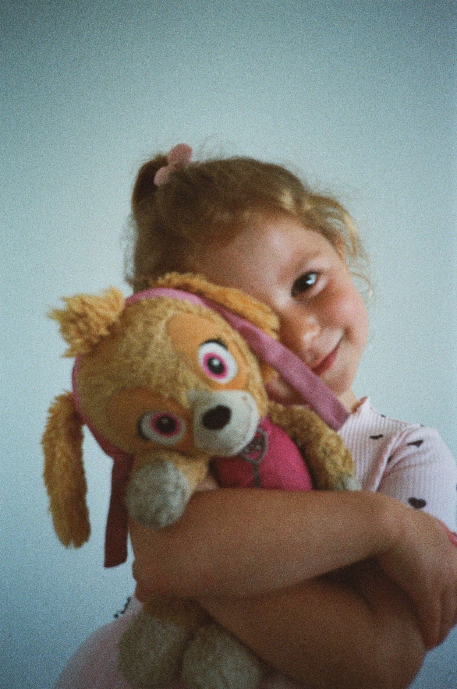 A smiling young girl with curly hair hugs a Skye PAW Patrol stuffed animal toy against a light background.