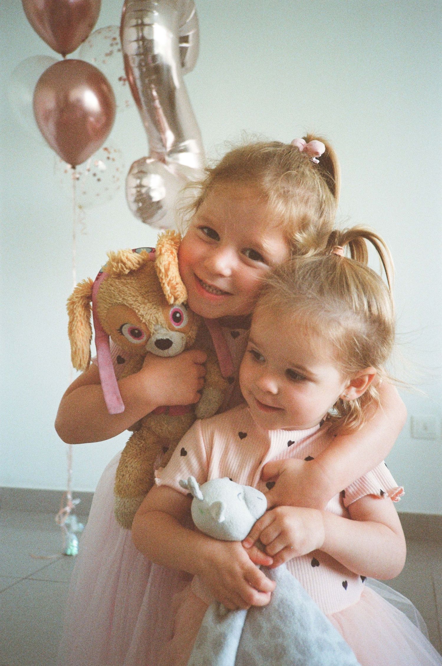 Two young girls in pink tutu dresses hugging each other while holding stuffed animals at a birthday party with balloons.
