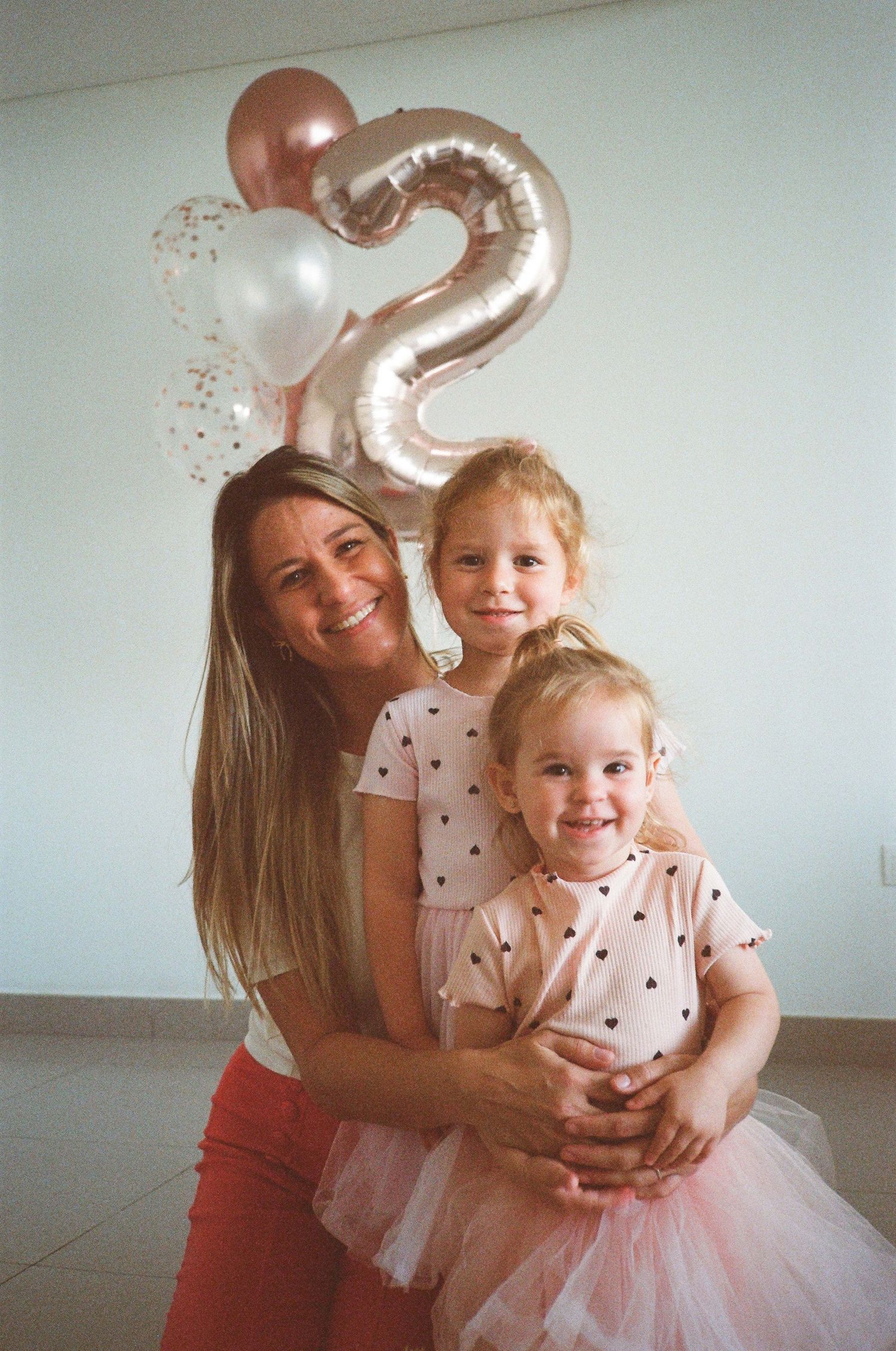 A smiling woman hugs two young girls in matching outfits at a 2nd birthday party with balloons.