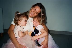 Smiling woman holding two toddler girls in pink tutus, one clutching a black and blue toy.