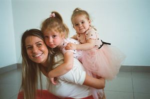 A smiling woman gives two young girls in pink tutus a piggyback ride indoors.