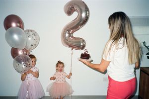 Two young girls in pink tutus celebrate their 2nd birthday with balloons as a woman brings a cake.