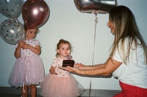 Two young girls in pink tutu skirts celebrate a birthday as a woman presents a chocolate cake with a candle.