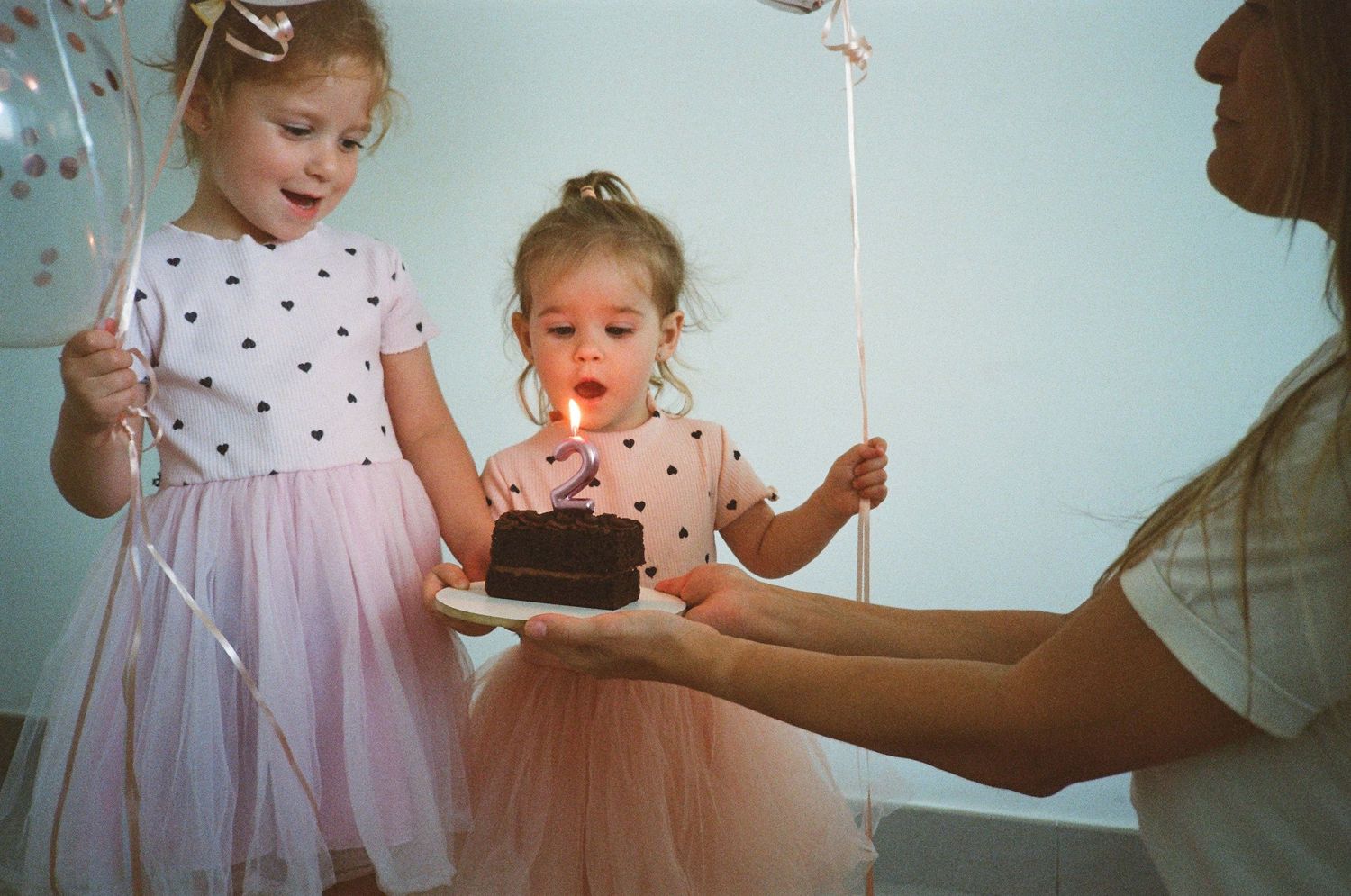 Two young girls in pink tutu skirts celebrate a birthday with a chocolate cake and lit candle held by an adult.