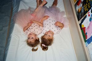 Two young girls in pink tutus lying on a white bed, smiling up at the camera in a playful pose.