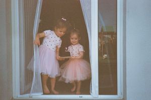 Two young girls in matching pink tutu skirts and polka dot tops standing by a white window.