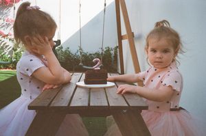 Two toddler girls in pink tutus sit at a wooden picnic table with a birthday cake showing number 2 candle outdoors.