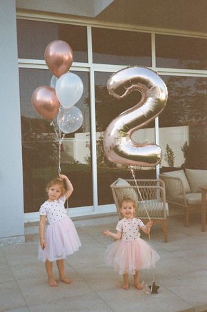 Two toddler girls in pink tutu dresses hold balloons and a number 2 balloon celebrating their 2nd birthday.