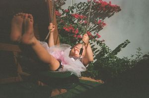 A young girl in a pink tutu relaxes on a wooden swing surrounded by vibrant red flowering bushes.