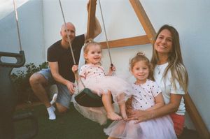 A happy family of four with two young girls in pink tutus sitting on a rope swing outdoors.