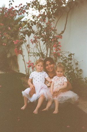 A woman sits outdoors with two young girls in matching white polka dot tutus, surrounded by vibrant pink bougainvillea.