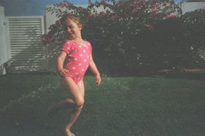 Young girl in pink polka dot swimsuit playing in sprinkler on green lawn during summer day.