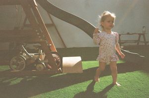 Young toddler girl in floral dress playing on green artificial grass near a playground slide in bright sunlight.
