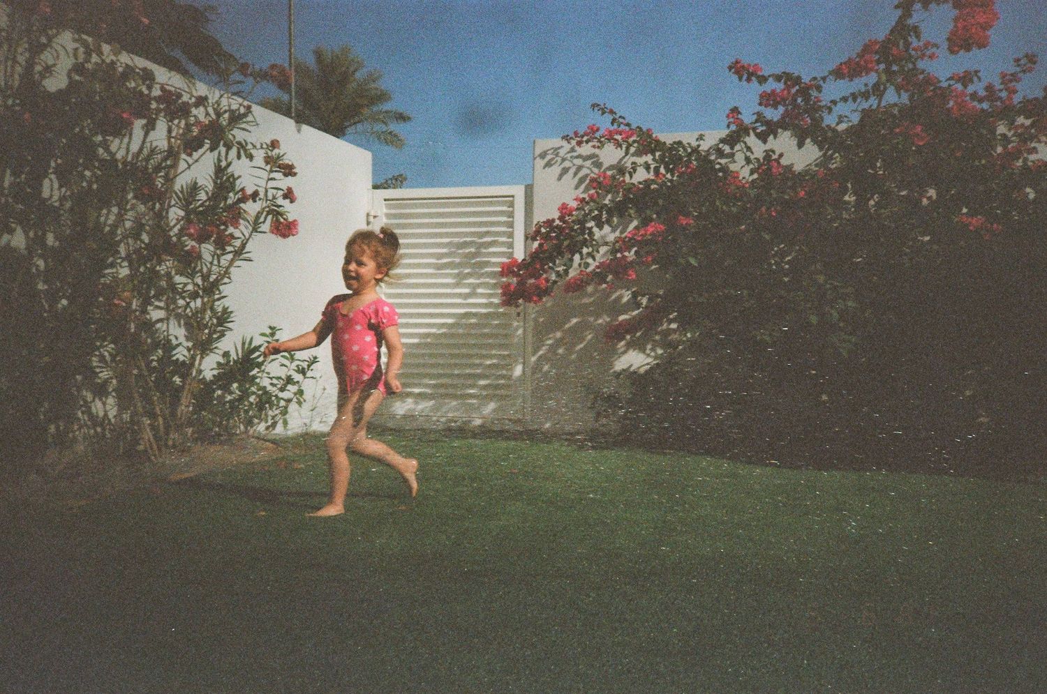 Young girl in pink swimsuit running on green grass in a sunny backyard with white walls and flowering bushes.