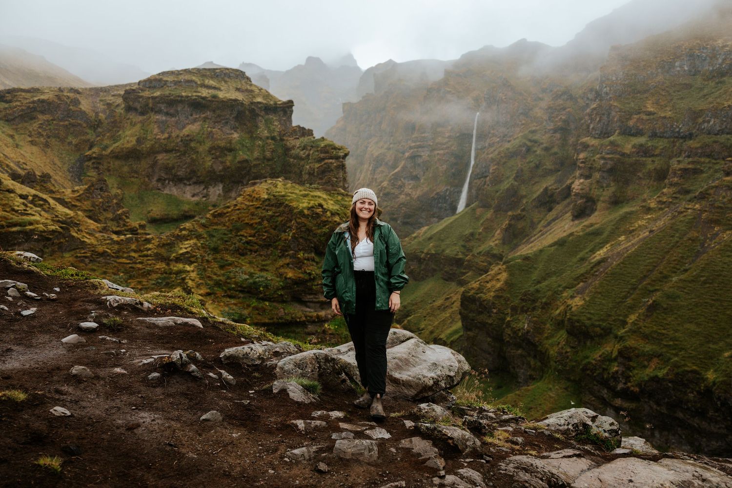 girl standing at Mulagljufur Canyon, Iceland with a view of the canyon and waterfall behind her on a rainy day